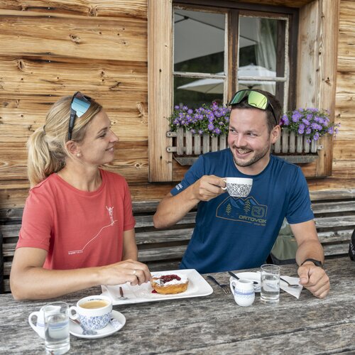 Two cyclists sit at a rustic wooden table with helmets nearby.