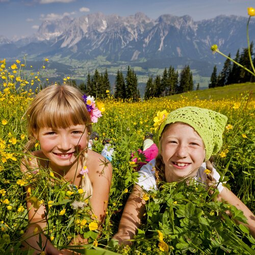 Two smiling girls lie in a yellow flower field with mountains.