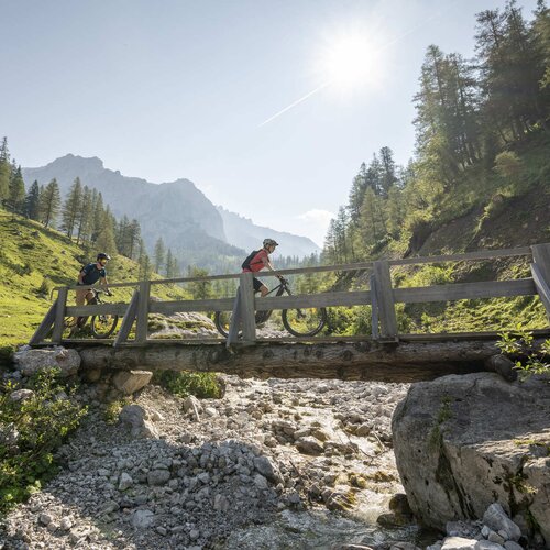 Two cyclists on a wooden bridge over a rocky stream.