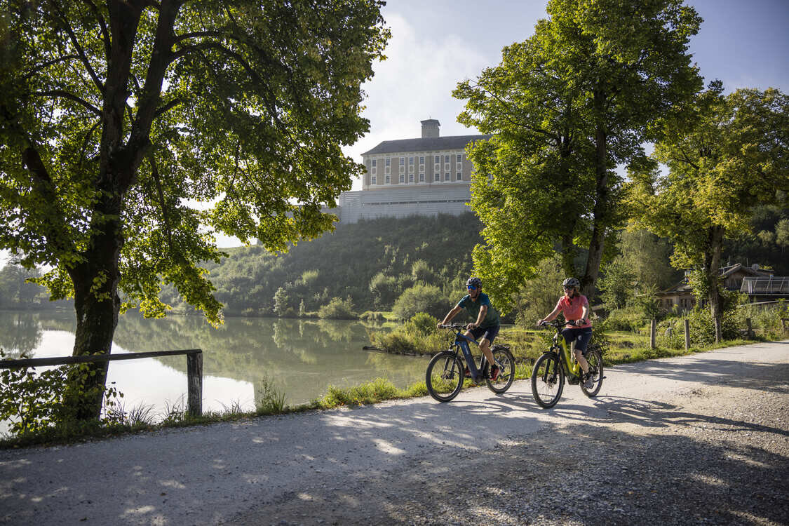 Two cyclists ride along a lakeside path framed by trees