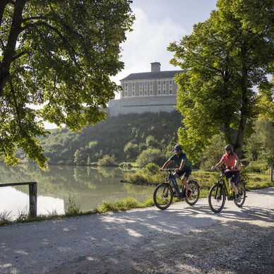 Two cyclists ride along a lakeside path framed by trees