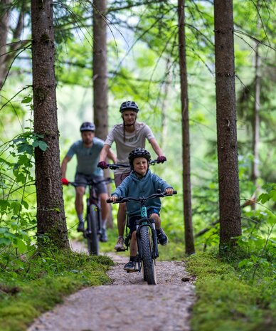Young child rides a bike on forest path with two adults following