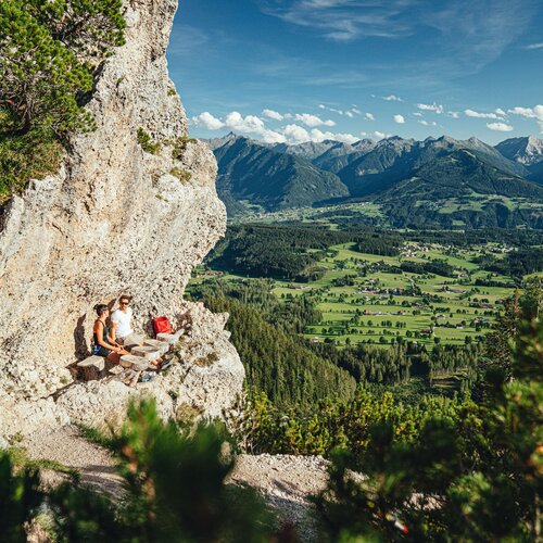 Two hikers sit on rocky cliff overlooking valley and mountains