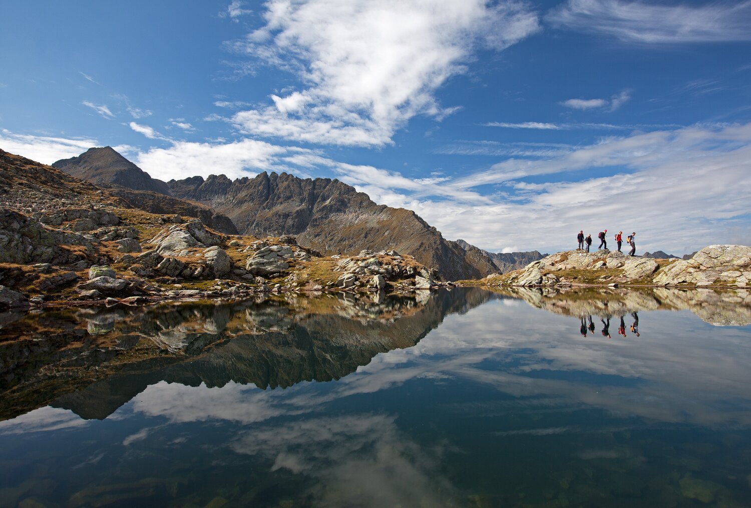 Group of hikers on rocky ridge above calm lake, mountains