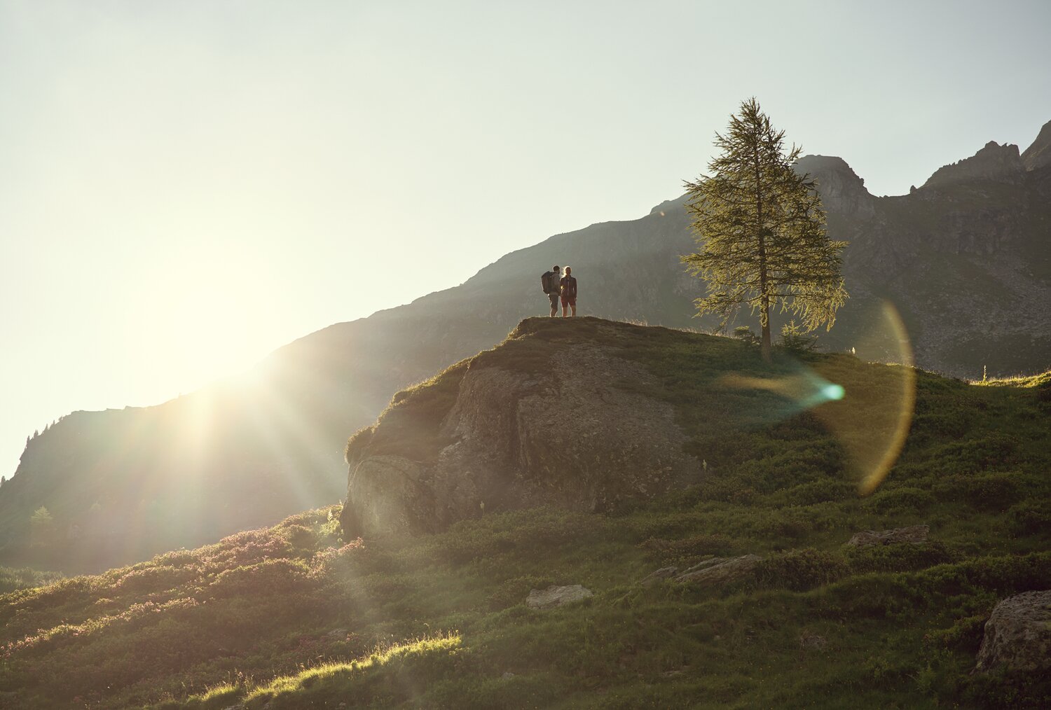 Two hikers stand on a rocky hilltop at sunset with mountains