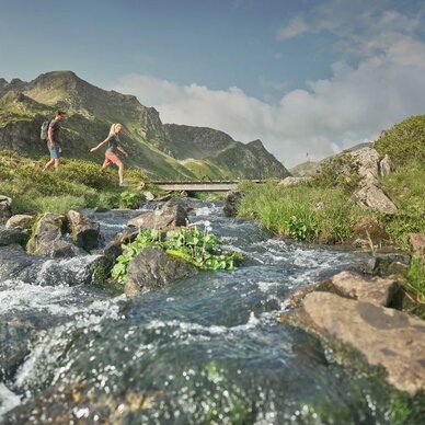 Two hikers cross a rocky stream in a mountain landscape.