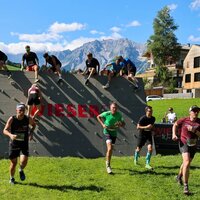 Runners sprint across a grass field beside a climbing wall obstacle