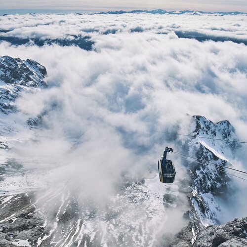 Cable car suspended above snow-covered mountains and clouds. | © Robert Maybach