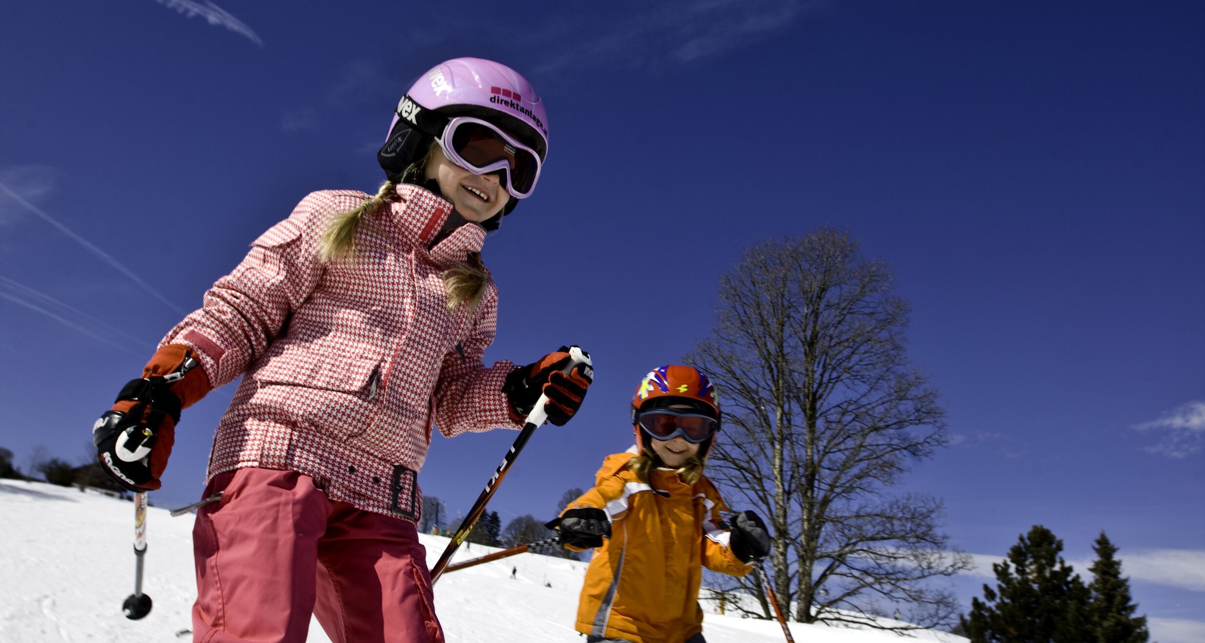 Two children skiing down a snowy slope under a clear blue sky. | © Tom Lamm