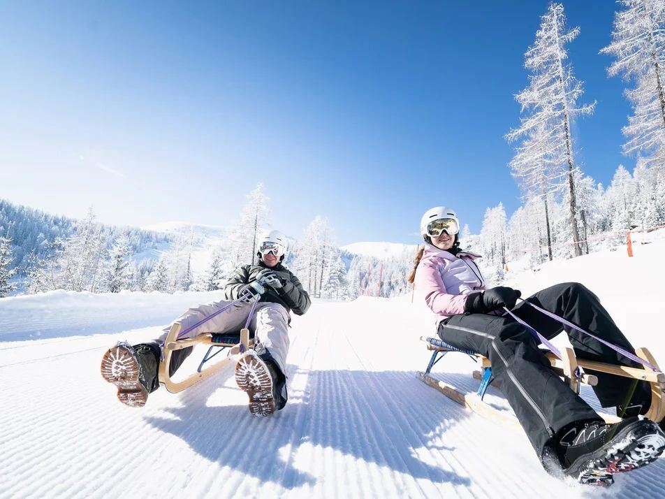 Two people sledding on wooden toboggans down a snowy slope