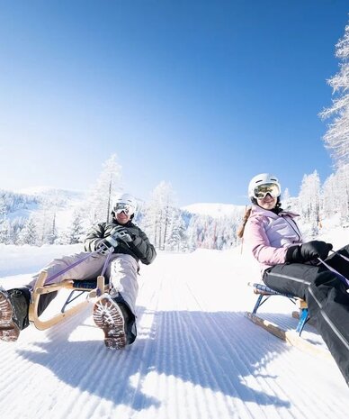 Two people sledding on wooden toboggans down a snowy slope