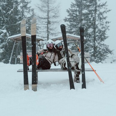 Two skiers sitting on a bench in the snow with skis nearby.