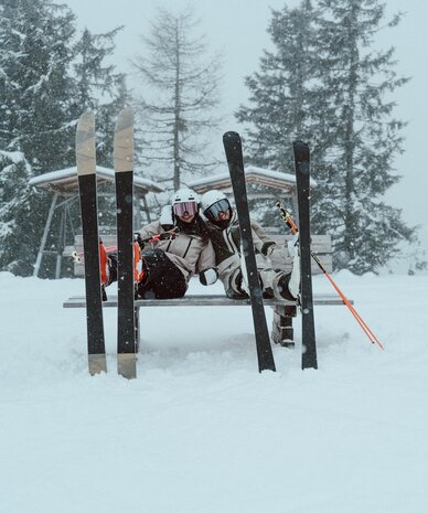 Two skiers sitting on a bench in the snow with skis nearby.