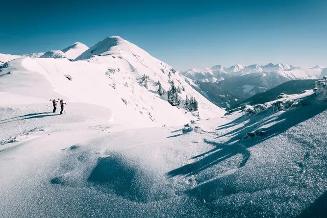 Two hikers standing on snowy mountain ridge under clear blue sky.