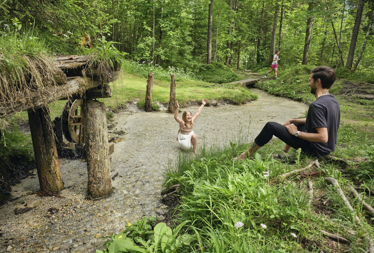 Girl splashes in shallow stream while man sits nearby. | © RAPHAELGABAUER.COM