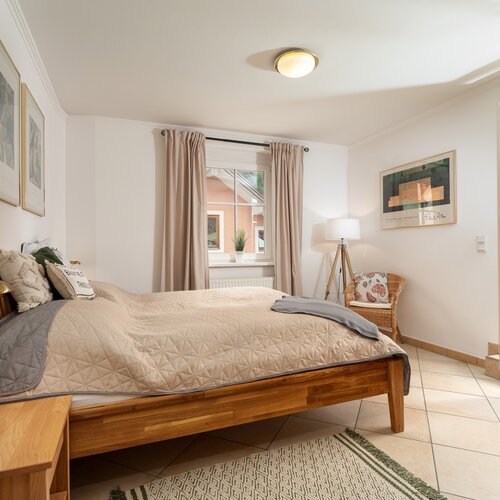 Schlafzimmer mit Holzbett, beigefarbener Steppdecke und Korbsessel.