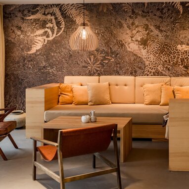 Cozy lounge area with beige sectional, wooden chairs, and a jungle mural.