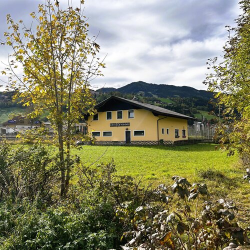 Vordergrund Bäume rahmen gelbes Haus in hügeliger Landschaft