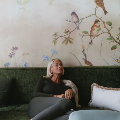Older woman resting on green sofa with floral bird mural behind.